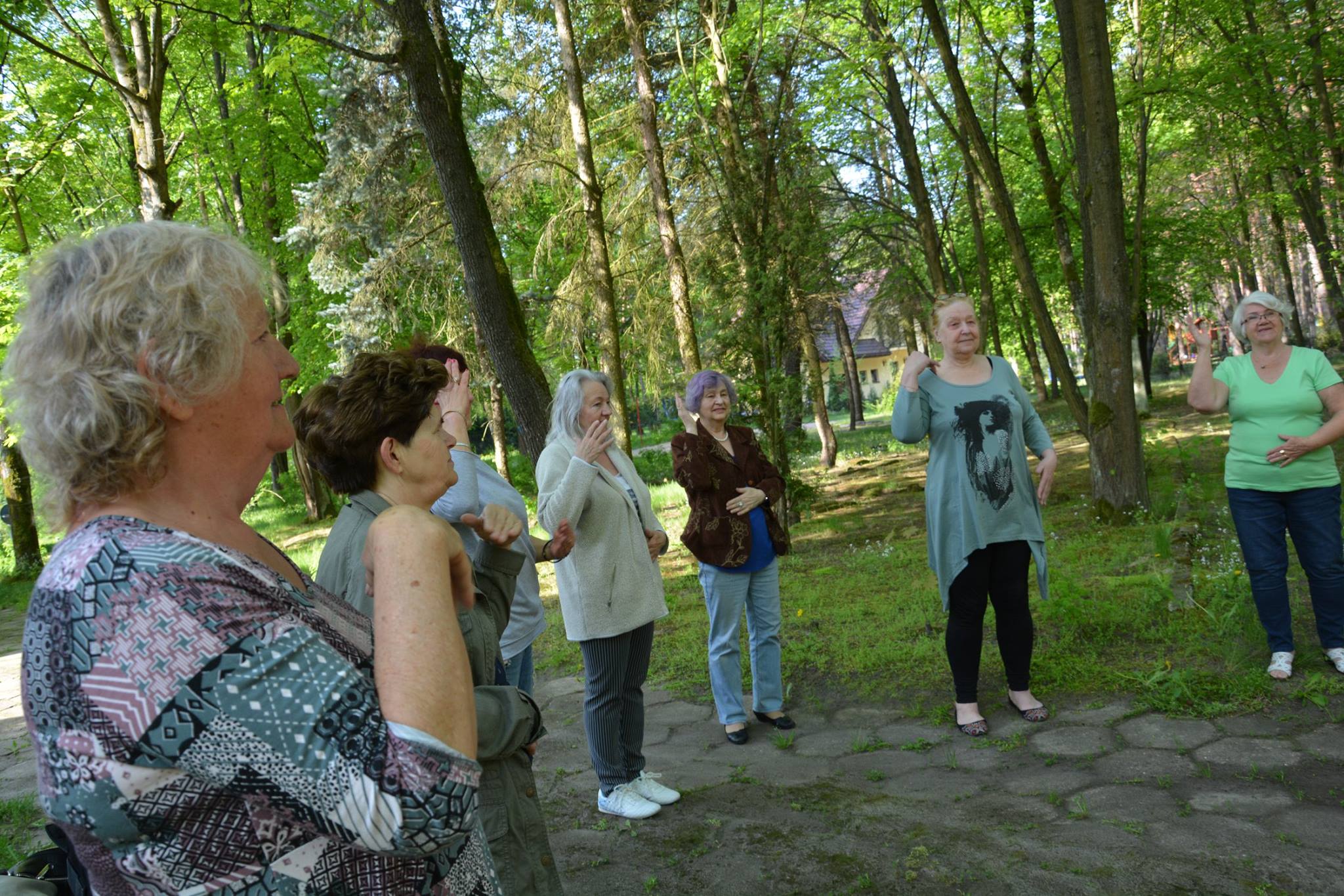 Zdjęcie przedstawia seniorki stojące w pół-okręgu, wykonujące jakieś ćwiczenie, wszystkie podnoszą jedną z rąk na wysokość ramienia.