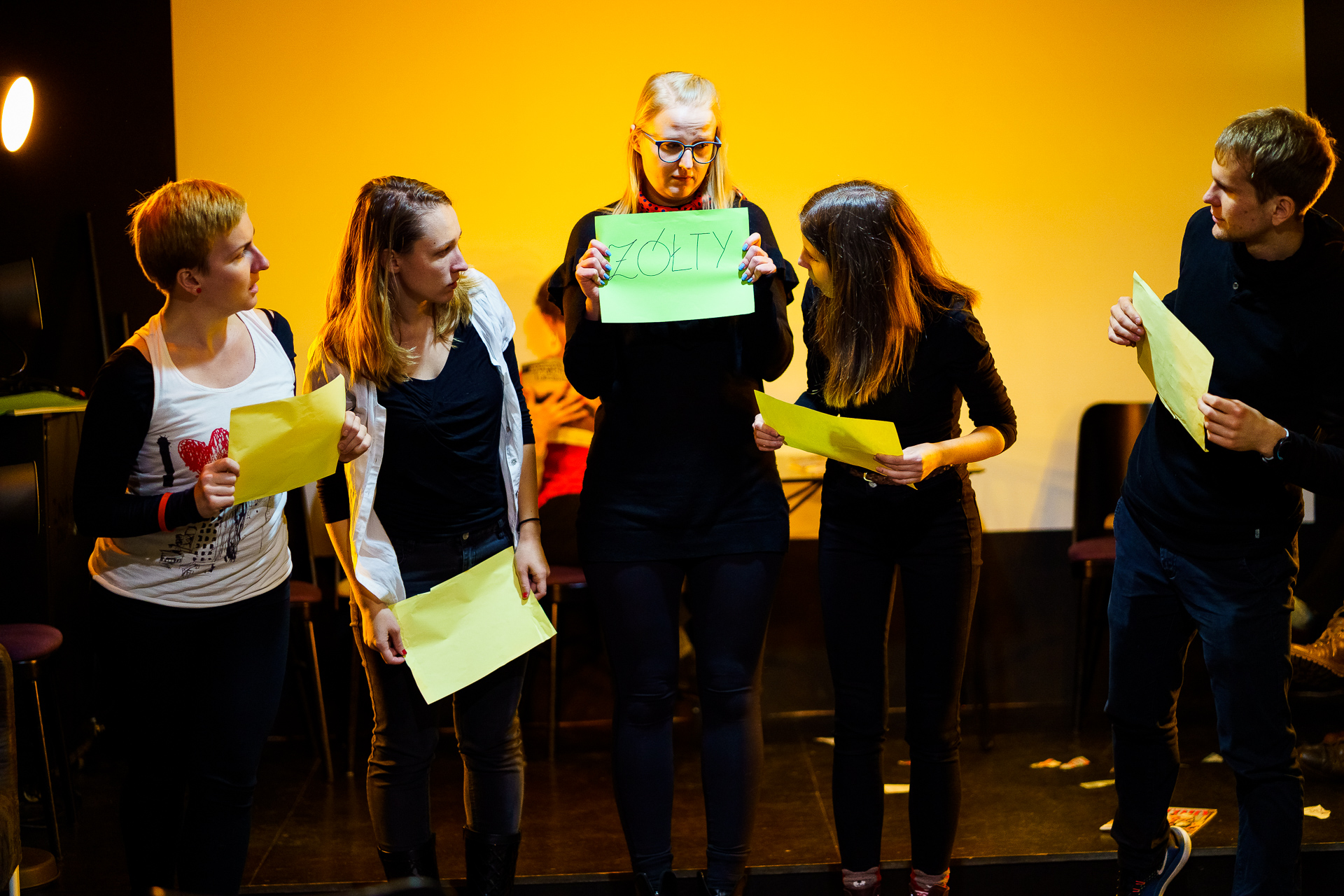 Zdjęcie wykonane podczas spektaklu teatru Gazetowego, przedstawia piątkę osób aktorskich na scenie. Jedna osoba trzyma zieloną kartkę z napisem "żółty" pozostałe osoby trzymają żółte kartki i patrzą ze zdziwieniem na osobę trzymającą zieloną kartkę