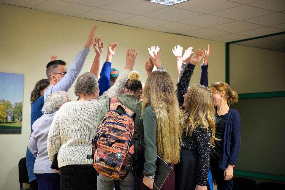 Zdjęcie przedstawia grupę osób, które stoją w kręgu blisko siedzie i każda z nich ma podniesioną jedną rękę do góry. Za zdjęciu widzimy osoby stojące plecami do osoby robiącej zdjęcie.