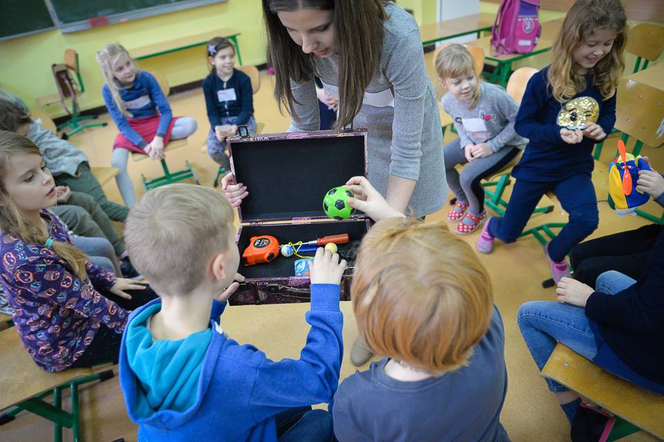 Na centralnym planie zdjęcia widać trenerkę trzymającą otwartą niewielką walizkę w której znajdują się różne przedmioty jedno z dzieci wyciąga małą piłeczkę, drugie wybiera co weźmie. W tle widać dzieci siedzące w kręgu