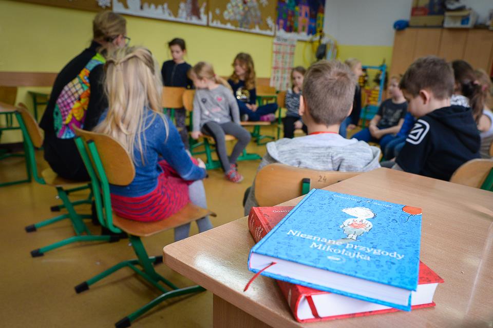 W tle zdjęcia widać dzieci siedzące w kręgu podczas warsztatów a w centralnym miejscu zdjęcia widać książkę Nieznane przygody Mikołajka