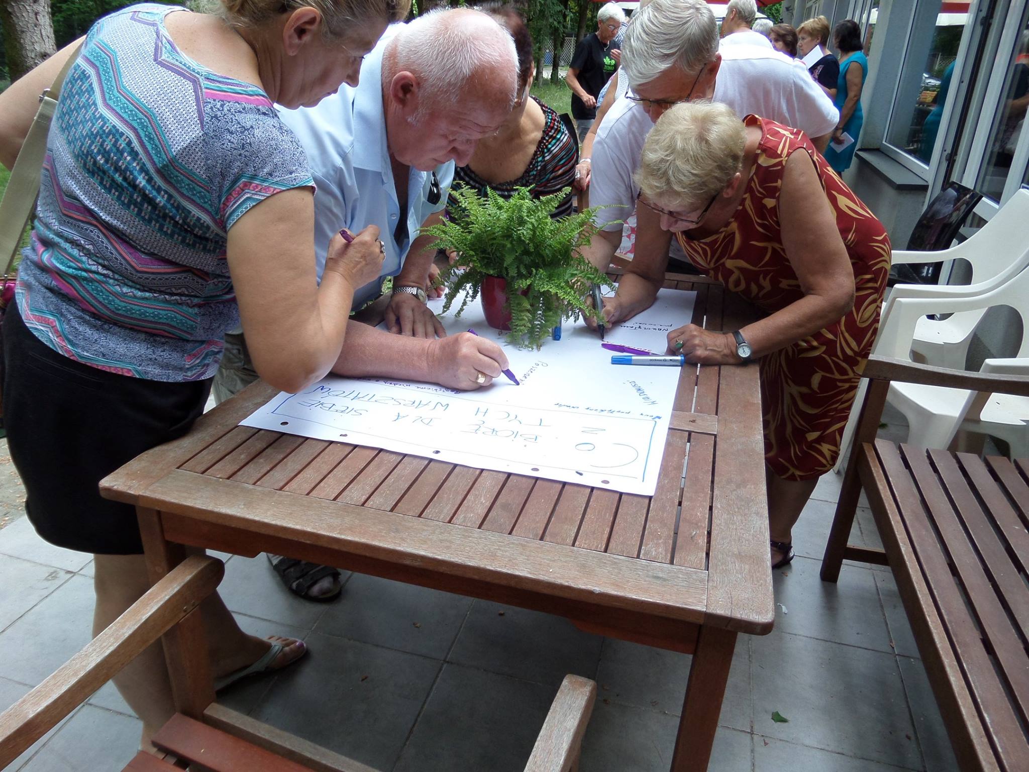 STI_3 Zdjęcie przedstawia 5 osób seniorskich pochylające się i piszące coś na dużej kartce typu flipchart