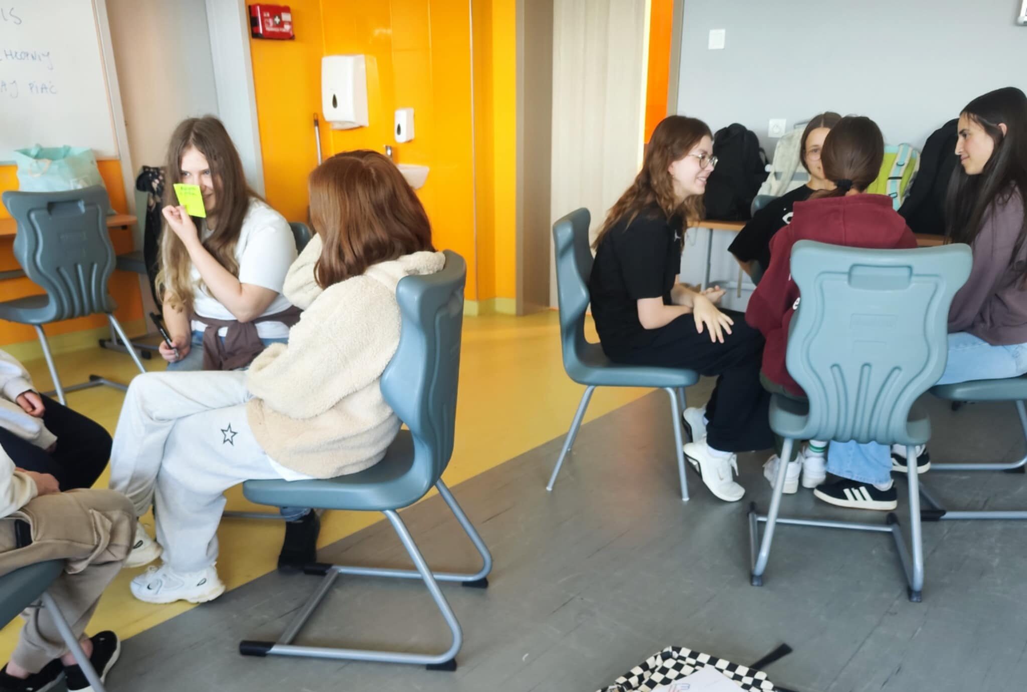 Ósemka uczniów i uczennic siedzących na krzesłach i pracujących w podgrupach. Leżący na podłodze plecak Janka (bohatera historii) oraz arkusz papieru z narysowaną sylwetką.